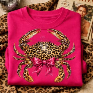 May include: A hot pink sweatshirt featuring a detailed crab design. The crab has a leopard print pattern on its body and claws, with a pink bow. The sweatshirt is folded on a leopard print fabric.