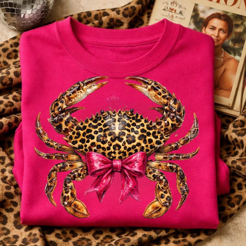 May include: A hot pink sweatshirt featuring a detailed crab design. The crab has a leopard print pattern on its body and claws, with a pink bow. The sweatshirt is folded on a leopard print fabric.