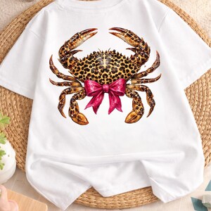 May include: White t-shirt featuring a detailed crab illustration. The crab has a leopard print pattern on its shell and claws, with a bright pink bow. The shirt is made of a soft, comfortable material.