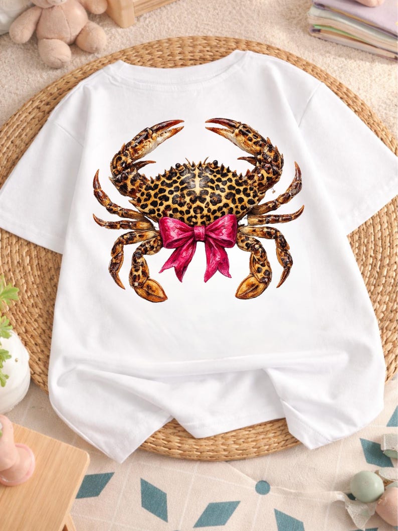 May include: White t-shirt featuring a detailed crab illustration. The crab has a leopard print pattern on its shell and claws, with a bright pink bow. The shirt is made of a soft, comfortable material.