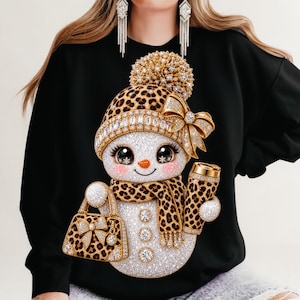 May include: A black long-sleeve sweatshirt featuring a graphic of a sparkling snowman wearing a leopard print hat with a gold bow and a matching leopard print scarf. The snowman holds a leopard print handbag and a tumbler, accented with crystals.