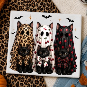 May include: A rectangular white cutting board featuring an illustration of three black kittens wearing ghost costumes. The ghosts have leopard print, cherry, and red polka dot patterns. Each kitten wears a matching bow tie, with bats and stars.