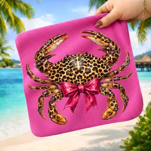 May include: A pink square towel featuring a detailed crab illustration with a leopard print pattern on its shell and legs. The crab has a bright pink bow. The towel is held up by a hand, with a beach scene in the background.