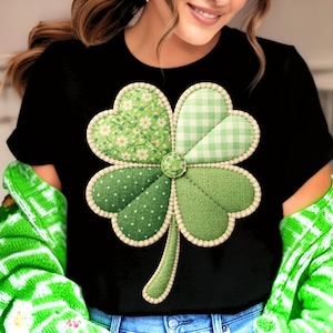 May include: Black t-shirt featuring a large, detailed shamrock design. The shamrock is composed of four green heart-shaped leaves, each with a different pattern: floral, gingham, polka dots, and solid. The design is outlined with a cream-colored border.