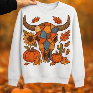 Puede incluir: Sudadera blanca con un diseño de calavera de toro de retazos coloridos. El diseño incluye calabazas, girasoles, hojas y un cactus, todo en colores otoñales. La sudadera es sostenida por una persona.