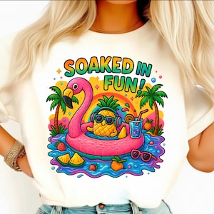 May include: White t-shirt with a colorful graphic. The graphic features a pineapple wearing sunglasses and headphones, floating on a pink flamingo float in water. The text "SOAKED IN FUN!" is above the image, with palm trees and a rainbow in the background.