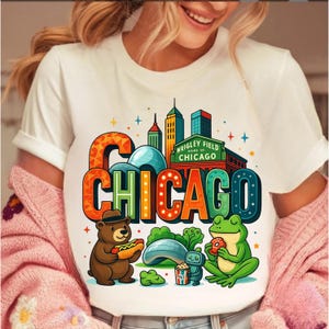 May include: White t-shirt with a colourful graphic of Chicago landmarks and cartoon characters. The design includes the word "CHICAGO", a bear with a hot dog, and a frog with a donut.