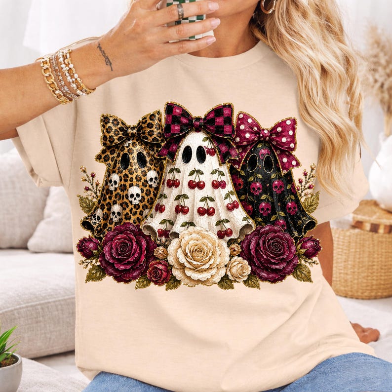 May include: A beige t-shirt featuring a graphic of three ghosts wearing large bows. The left ghost has a leopard print with skulls, the center ghost has a cherry pattern, and the right ghost has a skull pattern. Below are red and white roses.