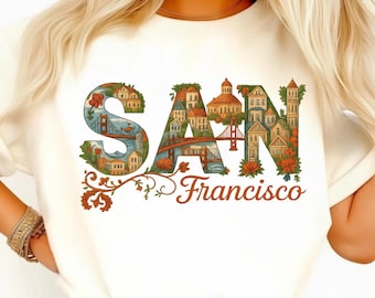 Puede incluir: Camiseta color crema con las palabras "SAN" y "Francisco" en un diseño decorativo. Las letras están llenas de ilustraciones de casas, puentes y flores. El diseño es en tonos de azul, naranja y verde.