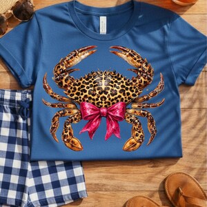 May include: A blue t-shirt featuring a crab design with a leopard print body and a pink bow. The crab's claws are a mix of brown and orange. The shirt is paired with blue and white checkered shorts and brown sandals.