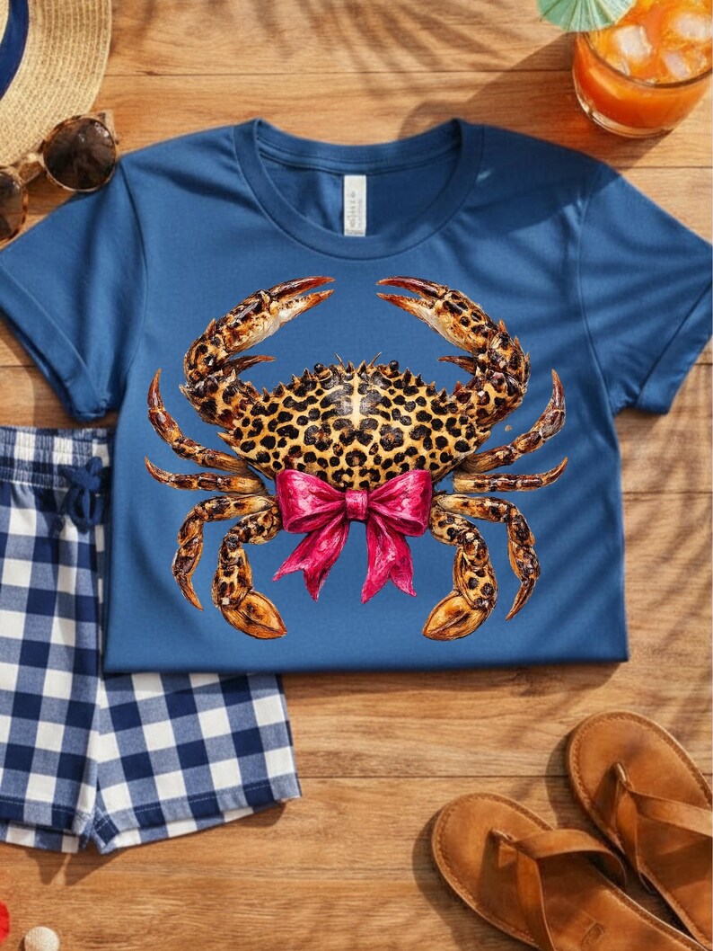 May include: A blue t-shirt featuring a crab design with a leopard print body and a pink bow. The crab's claws are a mix of brown and orange. The shirt is paired with blue and white checkered shorts and brown sandals.
