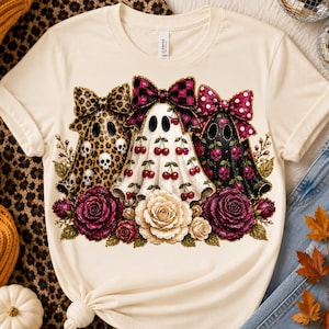 May include: A cream-colored t-shirt featuring a graphic of three ghosts wearing large bows. The left ghost has a leopard print with skulls, the center ghost has a cherry pattern, and the right ghost has a black cherry and skull print. Deep red and cream roses are at the base.