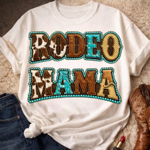 May include: White t-shirt with the words "RODEO MAMA" in a western-style design. The letters are brown, turquoise, and gold, with a cowhide pattern and turquoise bead trim. A pair of blue jeans and cowboy boots are visible.