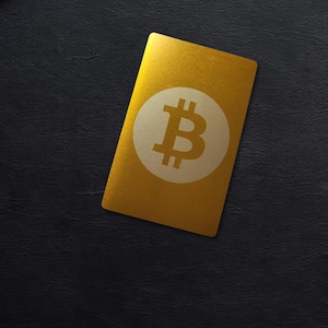 Op de afbeelding: Een goudkleurige kaart met een Bitcoin-symbool, geplaatst op een zwart oppervlak in de buurt van een laptop, notitieboek en tablet. Het Bitcoin-logo is een witte 'B' met verticale lijnen. De kaart is rechthoekig.