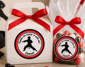 Op de afbeelding: Een witte kartonnen doos met handvat en een doorzichtig plastic zakje gevuld met rode, witte en zwarte snoepjes. Beide zijn voorzien van een ronde sticker met een zwarte silhouet van een vechtsporter en de tekst "Thanks for kickin' it with me! Samuel". Rode satijnen linten zijn bovenop gestrikt.