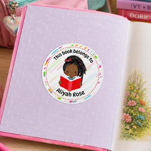 May include: A circular sticker on the inside cover of a book featuring a cartoon illustration of a child with dark skin and braided hair reading a red book. The text reads This book belongs to Aliyah Rose, surrounded by a colorful pastel striped border.