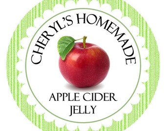 Op de afbeelding: Een rond etiket met een geschulpte rand en een lichtgroen verticaal gestreept patroon. De tekst CHERYL'S HOMEMADE staat gebogen boven een helderrode appel met een groen blaadje, en de tekst APPLE CIDER JELLY staat eronder.