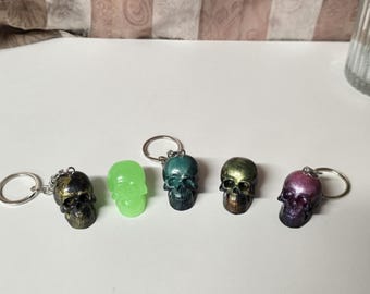 May include: Five skull-shaped keychains in a row. The skulls are in various colors, including black and gold, green, teal, gold, and purple. Each skull has a silver keyring attached.