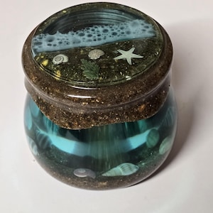 May include: A round glass jar with a lid featuring a beach scene. The lid has a sandy brown border and a clear top with a blue ocean and white foam design. The jar's interior is filled with blue liquid and seashells.