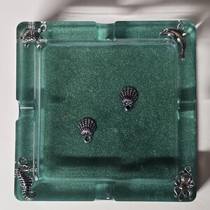May include: Square, sea-themed ashtray in shades of green. The ashtray features four silver charms: a turtle, a dolphin, a seahorse, and an octopus. Two silver seashell charms are in the center. The ashtray is made of a translucent material.