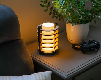 Op de afbeelding: Een cilindervormige houten lamp met een warme gloed, met een gelaagd ontwerp. De lamp staat op een grijze nachtkast naast een plant in een pot en een zwarte gamecontroller. Een gestreept kussen en een grijze fauteuil staan op de voorgrond.