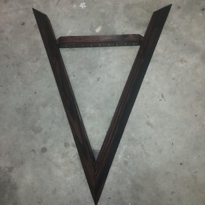 May include: Dark stained wooden V-shaped frame with a horizontal support piece. The frame has a pointed bottom and is set against a concrete surface. The wood has a rich, dark brown colour.