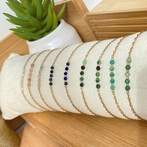 May include: A set of nine delicate gold chain bracelets with different coloured gemstone beads. The bracelets feature a gradient of colours, starting with light pink and transitioning to green.