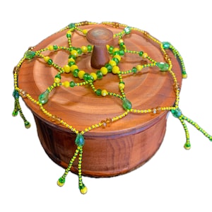 May include: A round wooden box with a lid, adorned with a beaded design in shades of yellow, green, and clear. The lid features a small, rounded wooden handle. The beaded design drapes over the box, creating a decorative effect.