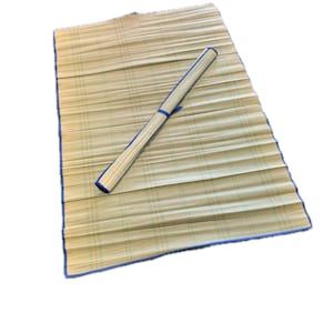 May include: A rectangular, woven mat with a blue border and a rolled-up section secured with a blue band. The mat is made of light-colored, natural fibers, and the rolled section is positioned diagonally across the mat.