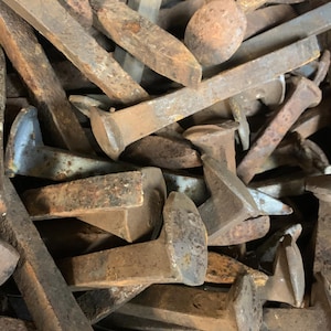 May include: A pile of weathered, rusty railroad spikes. The spikes are various sizes and shapes, with flat heads and tapered bodies. The metal has a brown and gray patina, indicating age and exposure to the elements.