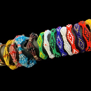 May include: A collection of beaded bracelets in various colors, including red, yellow, blue, green, and white. The bracelets are displayed on a black cylindrical stand against a black background. Some bracelets have intricate patterns.