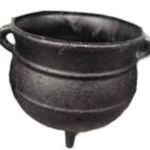 May include: A black cast iron cauldron with three short legs and two side handles. The cauldron has a rounded body and a wide opening. The exterior has a textured, slightly rough surface.