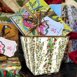 May include: A decorative white bin with a botanical pattern of green leaves and red berries. A gift box with a floral design and a tag that reads "Robin Raymond" is inside the bin. The bin has a gold rim.