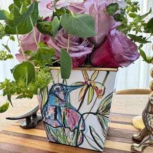 May include: A floral arrangement in a white square vase with a hummingbird design. The flowers are a mix of pink roses and other pink and white blooms, with green foliage. The vase sits on a wooden surface.