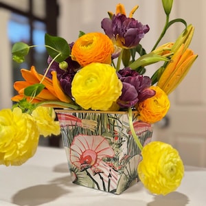May include: A vibrant floral arrangement in a decorative square container. The bouquet features yellow ranunculus, orange lilies, and deep purple tulips. The container has a whimsical print with mushrooms and foliage.