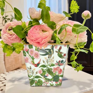 May include: A floral arrangement featuring pink flowers and green foliage in a decorative square vase. The vase is adorned with a hummingbird and floral pattern. The arrangement is placed on a white surface.