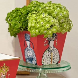 May include: A red, square planter with illustrations of figures in traditional attire, holding a large arrangement of green flowers. The planter sits on a clear glass cake stand.