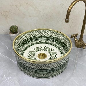 Puede incluir: Un lavabo redondo de cerámica con un patrón geométrico verde y blanco y un borde dorado. Un grifo de latón está posicionado a la derecha. Un pequeño cactus en maceta está a la izquierda. El lavabo es un accesorio decorativo para el baño.