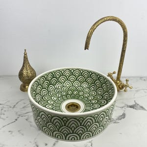 Puede incluir: Un lavabo redondo de cerámica con un diseño estampado en verde y blanco. El lavabo se combina con un grifo dorado y una linterna dorada decorativa. El diámetro del lavabo es de aproximadamente 40 cm.