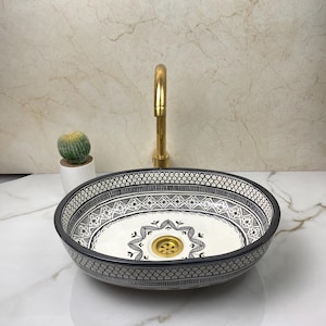 May include: Oval ceramic sink featuring a black and white geometric design. The sink has a gold-coloured drain and sits on a white marble countertop. A gold tap and a small cactus in a white pot are also visible.