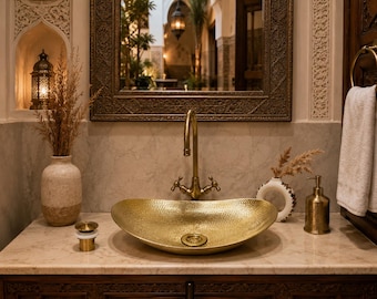 May include: A gold-toned, oval-shaped hammered metal vessel sink sits on a marble countertop. It features a tall, curved brass faucet with two handles. A matching gold drain plug, a textured beige vase with dried grass, and a soap dispenser are present.