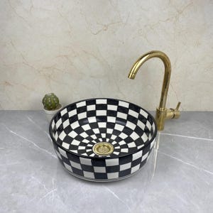 Op de afbeelding: Een zwart-wit geblokte keramische wastafel met een gouden kraan. De wastafel heeft een cirkelvormig ontwerp met een zwart-wit dambordpatroon. Links van de wastafel staat een kleine cactus in een witte pot.