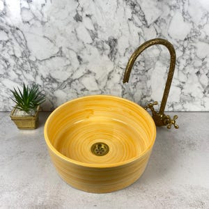 May include: A round, yellow ceramic vessel sink with a swirling, hand-painted finish. The basin features a central brass drain. Beside the sink stands a tall, curved, antique-style brass faucet with intricate etched detailing and cross handles.
