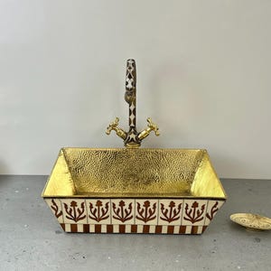 Könnte beinhalten: Ein goldfarbenes, gehämmertes Waschbecken aus Messing mit einem weißen und braunen Muster an der Kante. Das Waschbecken hat einen passenden goldfarbenen Wasserhahn mit einem weißen und braunen Muster am Griff.