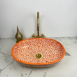 Puede incluir: Lavabo ovalado de cerámica con un patrón geométrico naranja y blanco. El lavabo tiene un grifo y un desagüe de latón. A la izquierda del lavabo hay una linterna decorativa de latón. El lavabo está sobre una encimera de mármol blanco.