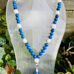 Op de afbeelding: Een blauwe en witte kralenketting met een grote blauwe traanvormige hanger. De ketting ligt op een witte displaystandaard.