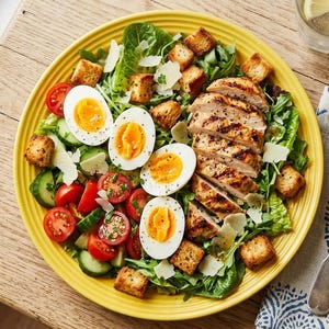 May include: A yellow plate containing a salad with sliced grilled chicken, halved hard-boiled eggs, cherry tomatoes, cucumber slices, croutons, and shaved cheese. A fork and knife rest on a patterned napkin beside the plate on a wooden surface.