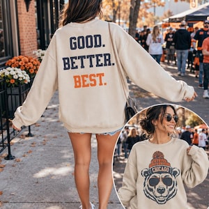 May include: A beige crewneck sweatshirt featuring the text GOOD. BETTER. BEST. in dark blue and orange on the back. The front displays a graphic of a bear wearing sunglasses and an orange pom-pom beanie with the word CHICAGO printed on the hat.