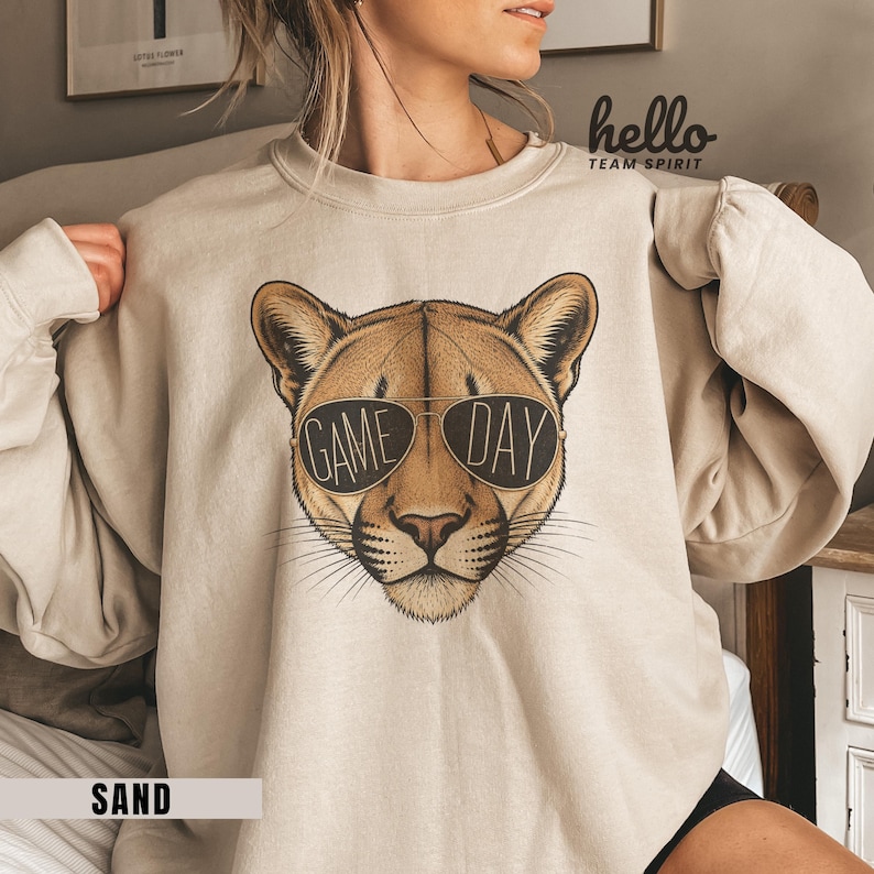 May include: Sand-colored sweatshirt featuring a cougar wearing sunglasses with the words "GAME DAY" printed on the lenses. The sweatshirt has long sleeves and a crew neck. The text "hello TEAM SPIRIT" is printed on the upper right side.