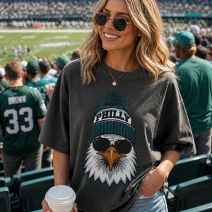 K&ouml;nnte beinhalten: Dunkelgraues T-Shirt mit einer Grafik eines Adlers, der eine Sonnenbrille und eine blaugr&uuml;ne und wei&szlig;e M&uuml;tze mit dem Wort "PHILLY" tr&auml;gt. Das Adlerdesign ist vor einem wei&szlig;en Hintergrund. Das Shirt wird mit Jeansshorts getragen.